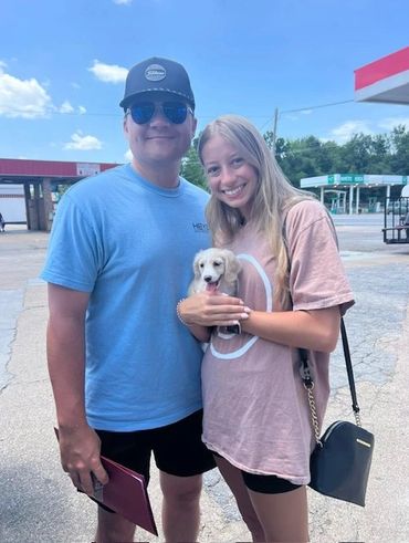 A smiling couple holding a small white puppy outdoors on a sunny day. long hair cream Dachshund puppy