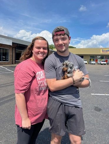 A couple standing outdoors with a small dog, smiling at the camera.