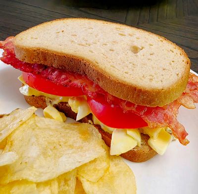 Bacon, tomato, and cheese sandwich with potato chips on a white plate.
