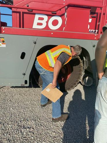 A worker inspects a large vehicle tire, holding a clipboard and wearing a safety vest.