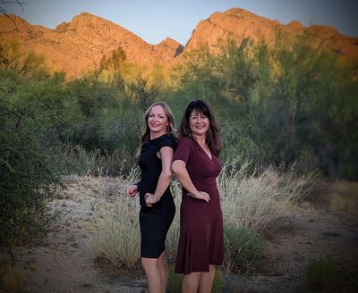 Two women posing back-to-back outdoors near mountains.