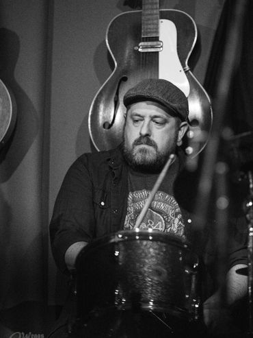 A bearded man playing drums with a guitar hanging on the wall behind him.