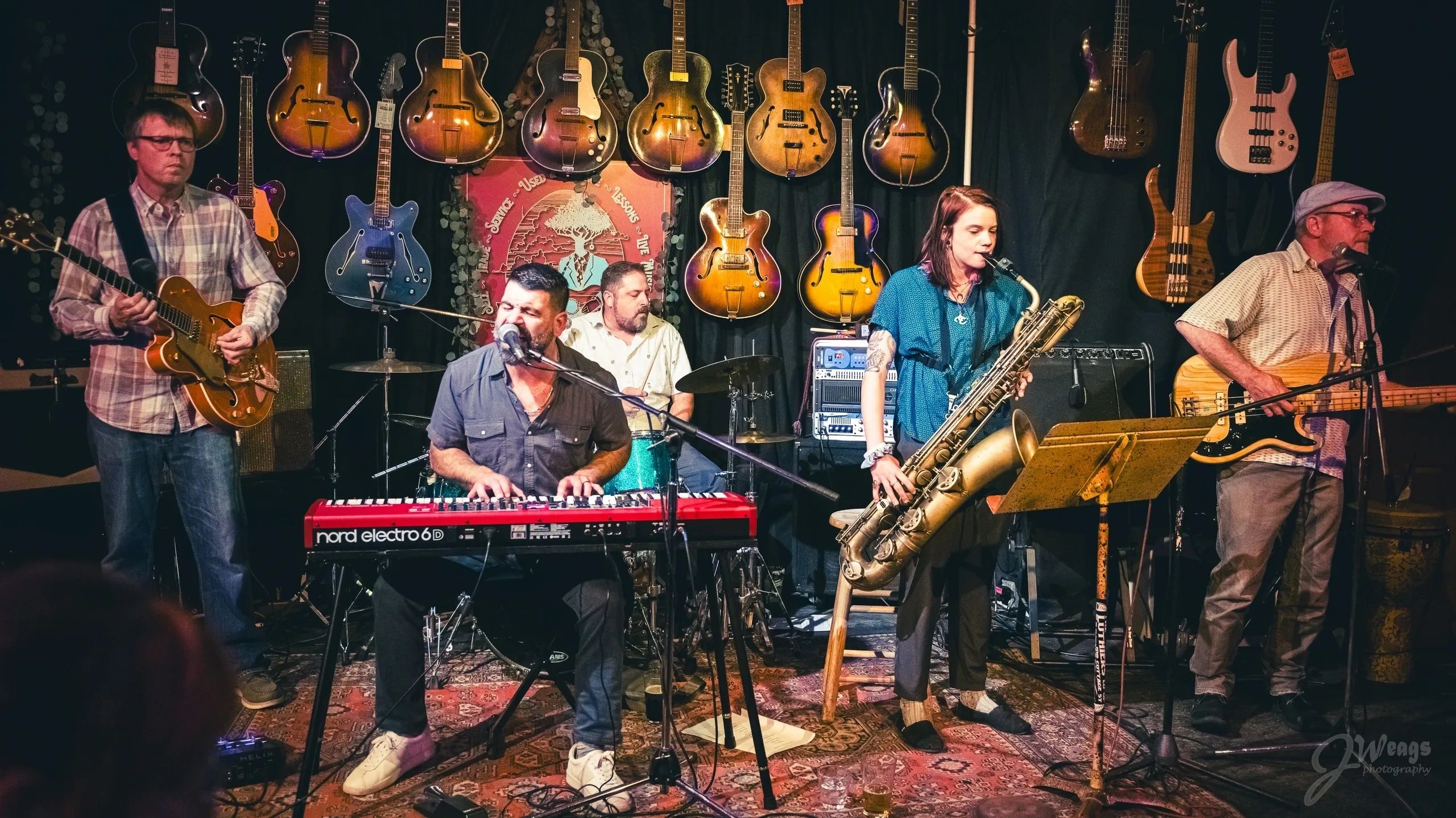 A band performs live with guitars, keyboard, saxophone, and drums in a cozy venue.
