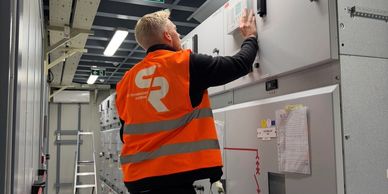 Factory Acceptance Testing Technician in orange vest working on electrical control panel inside an industrial facility.
