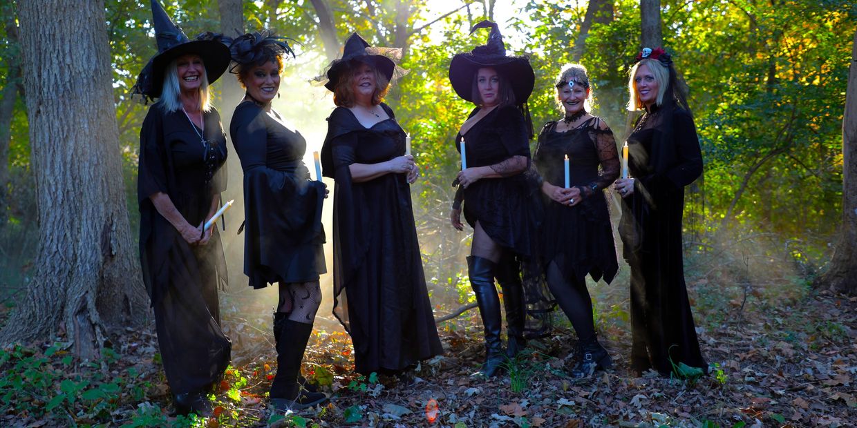 Six women dressed as witches holding candles in the woods.