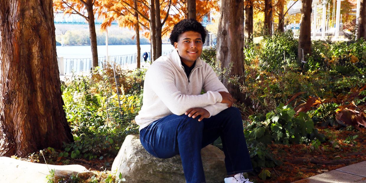 Young man in white sweater sitting outdoors near trees.