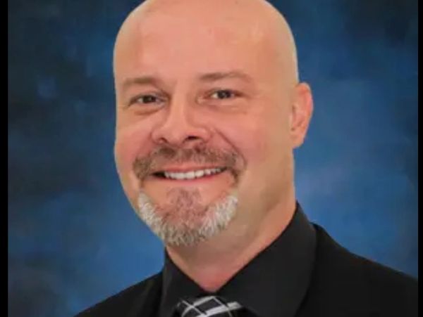Smiling bald man in a black shirt and patterned tie against a blue background.