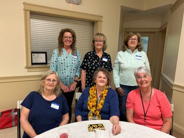 Group of six smiling women posing indoors around a table.