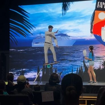 A man balances on a board on a roller while a woman watches on stage with a cruise ship backdrop.