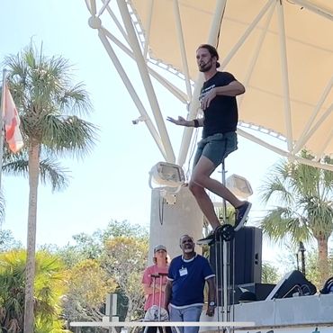 Man performing on a unicycle on an outdoor stage with an audience watching.