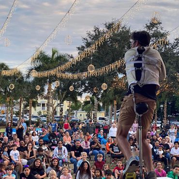 A performer on a unicycle entertains a large, diverse crowd at an outdoor event with festive lights.