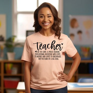Smiling woman wearing a light pink graphic t-shirt that says “Teacher: A multi-tasking educational rockstar who lives to inspire and loves to encourage” while standing in a colorful classroom setting.