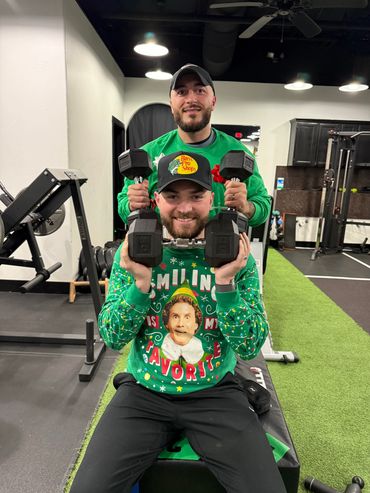 Two men in green sweaters holding dumbbells, posing in a gym.