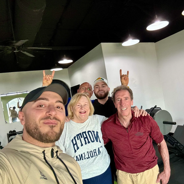 A group selfie at the gym with five happy people, two making playful hand signs.