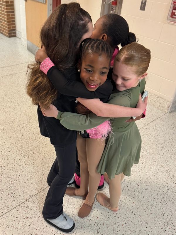 Four girls sharing a group hug in a hallway, dressed in dance attire.