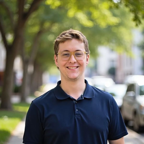 Young man smiling outdoors wearing glasses and a navy blue polo shirt.