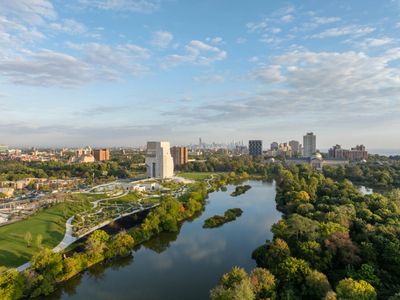 A river flowing through a city park with buildings in the background under a partly cloudy sky.