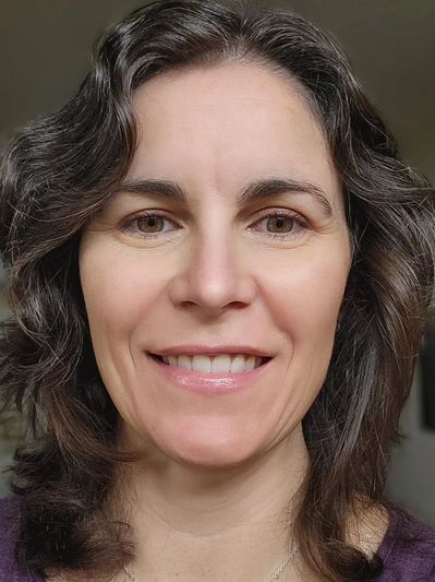 Smiling woman with wavy brown hair and light eyes wearing a purple top.