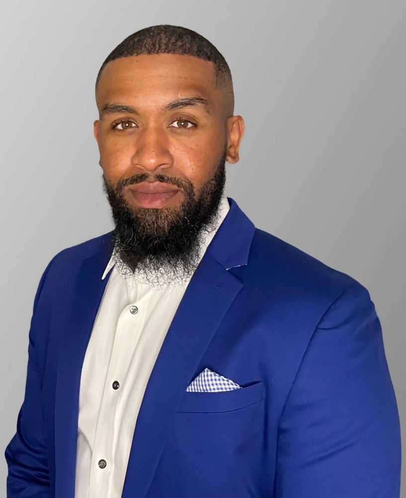 Man with beard wearing a blue suit and white shirt against a gray background.