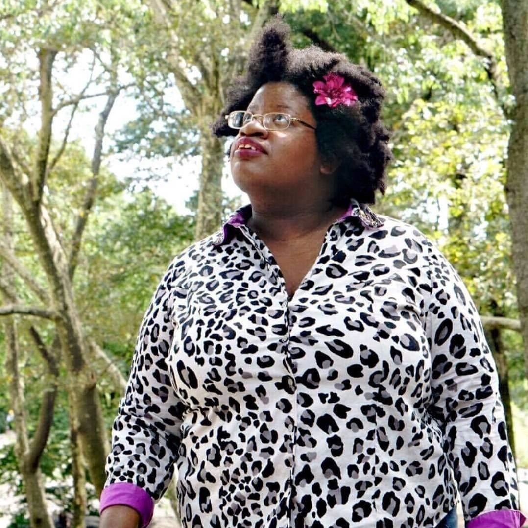 Pro photo of me, a proudly fat, brown-skinned, Black non-binary woman. Outdoors, day. Trees, sun.