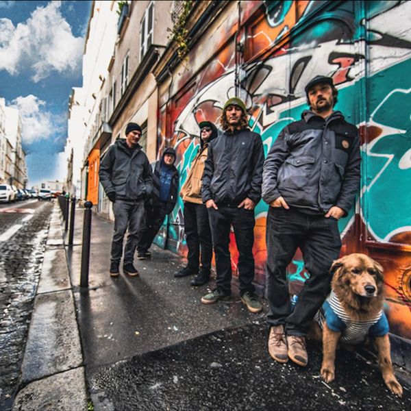 Five men and a dog standing by a graffiti-covered wall on a city street.