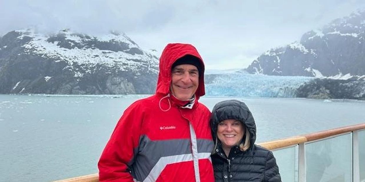 Couple in warm jackets smiling on a boat with snowy mountains and glacier behind.