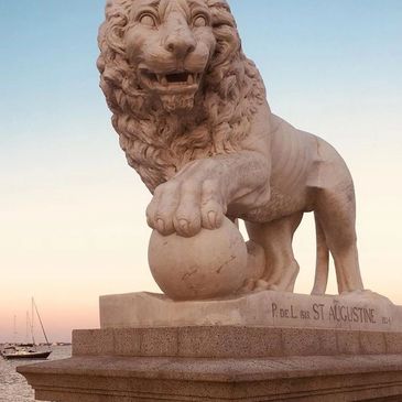 Stone lion statue with paw on a ball near a waterfront at sunset.