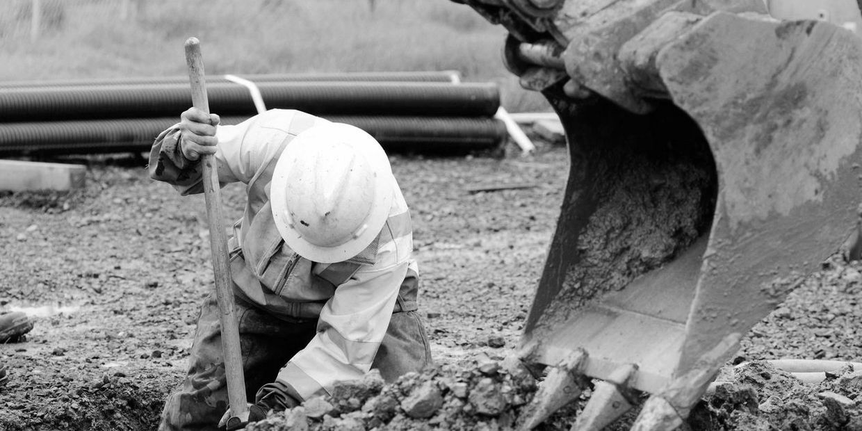 A person digging with a hand shovel, in a hole dug by an excavator.
