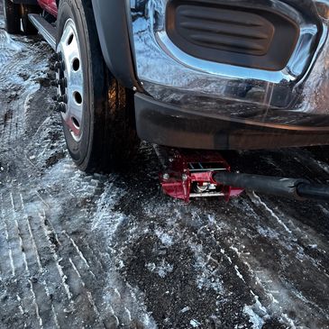 A car is lifted by a red hydraulic jack on a rough surface near a wall.