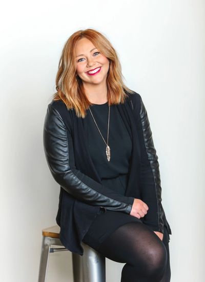 Smiling woman with red hair in black outfit sitting on a stool.