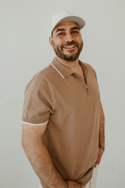 Smiling man in a beige polo shirt and white cap standing against a plain background.