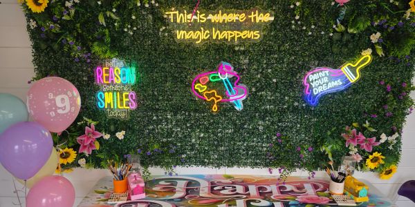 Colorful art-themed birthday setup with balloons and neon signs on a floral backdrop.