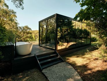Modern glass cabin with outdoor bathtub in a serene forest setting.