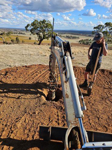 Man standing with a shovel next to an excavator on a sunny day in a rural area.