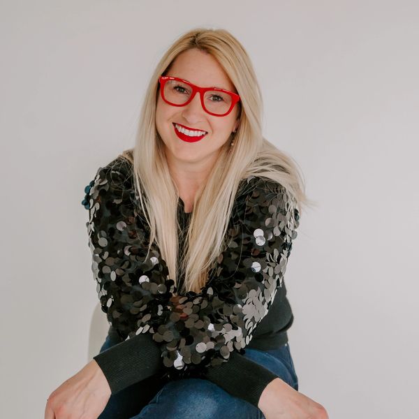 Blond woman smiling with red glasses and black sequin jacket.