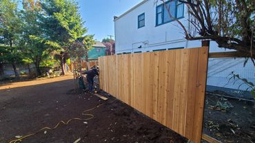 A person installing a new wooden fence beside a white building on a sunny day.