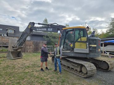 Two men shaking hands next to a Volvo excavator outside.