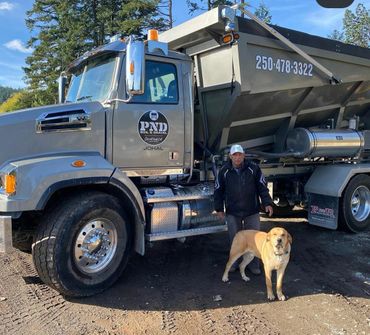 Man standing with a dog next to a large dump truck in a wooded area.