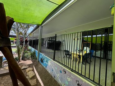 Outdoor school area with tables, chairs, and colorful mural beneath a green shade sail.
