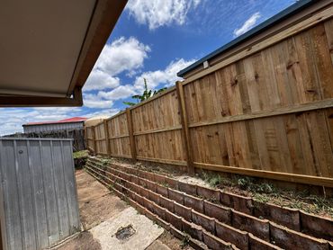 Backyard with wooden fence, retaining wall, and metal shed under a partly cloudy sky.