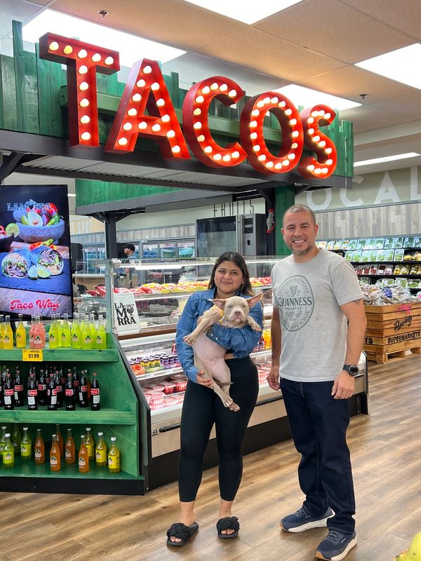 A smiling couple with a dog poses under a bright taco sign in a grocery store.