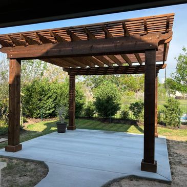 A wooden pergola stands on a concrete patio in a lush backyard.