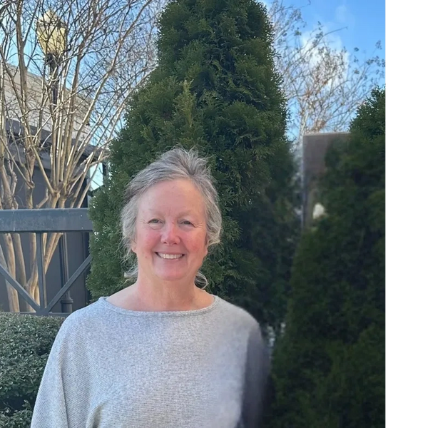 Smiling woman in a gray sweater stands outdoors beside green bushes.