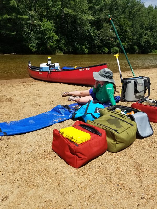 A child relaxes on a sandy riverbank beside a red canoe and outdoor gear.