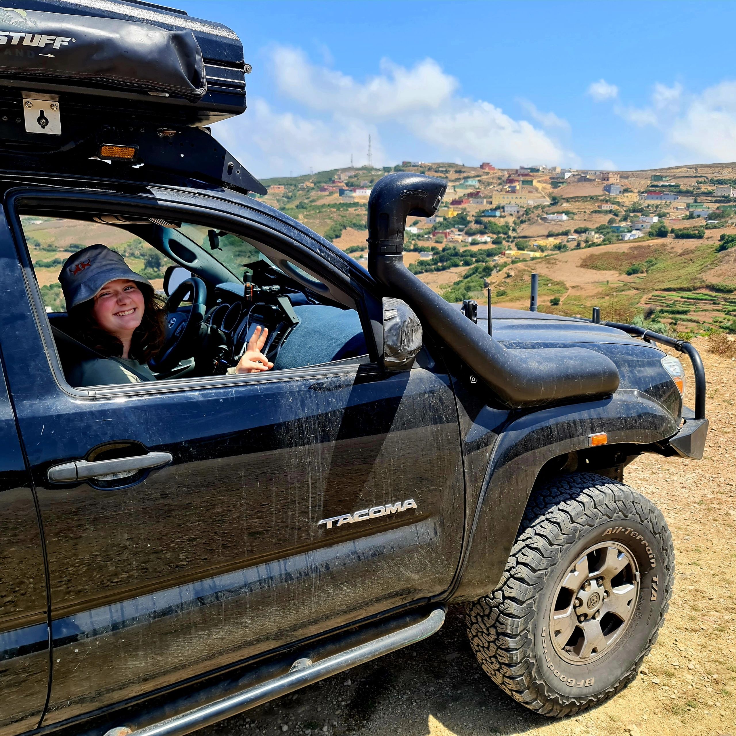 Smiling woman making peace sign in a black Toyota Tacoma with scenic hills in the background.
