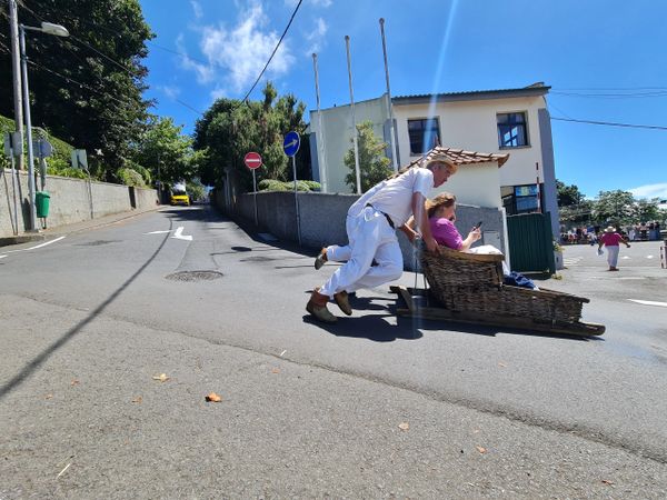 Man pushing woman in a wicker sled down a sunny street.