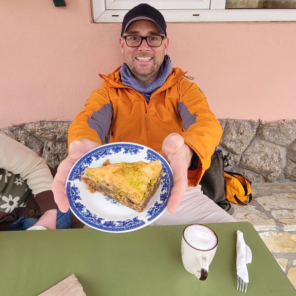 Smiling man in orange jacket offering a plate with baklava.