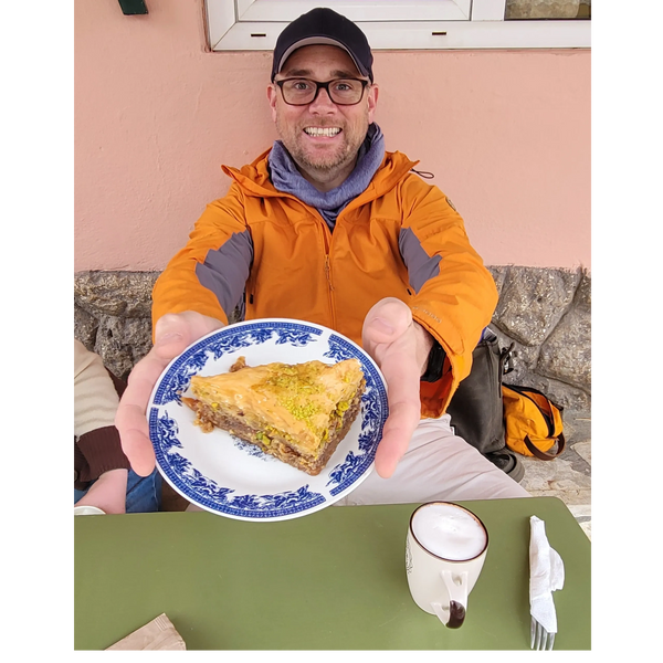 Man in orange jacket offering a plate with baklava dessert.
