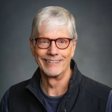 Smiling elderly man with glasses and gray hair against a dark background.