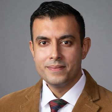 Professional headshot of a man in a brown jacket and patterned tie against gray background.